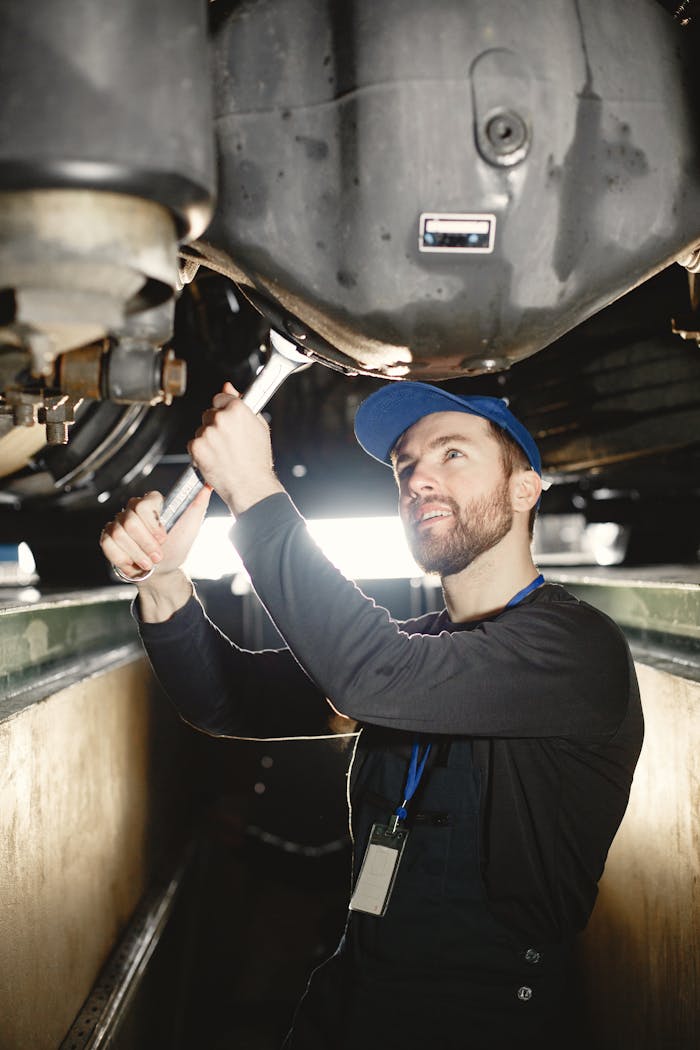 An adult male mechanic works on a vehicle's underside in a well-lit workshop.